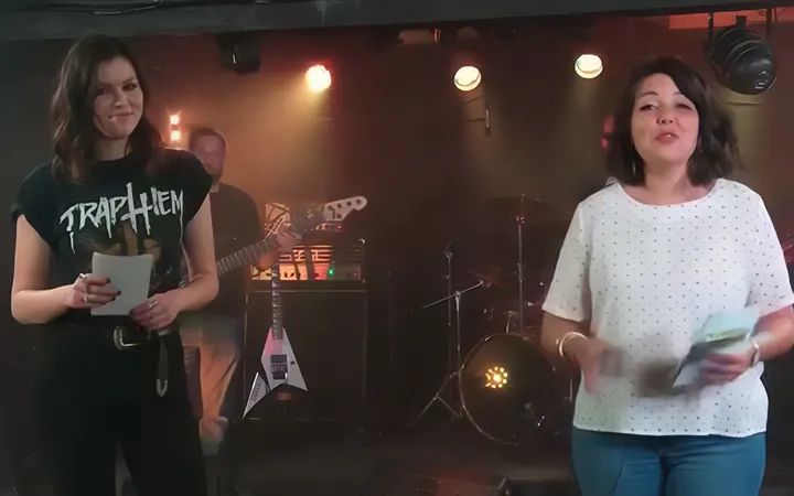 Une animatrice en haut blanc à pois s'exprime face caméra sur une scène de concert, guitares et batterie en arrière-plan